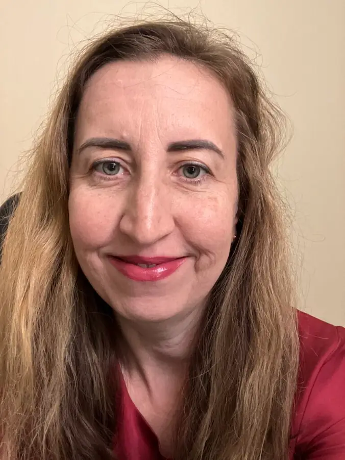 A close-up headshot of a smiling person with long, wavy light brown hair, wearing a reddish top against a plain beige background.