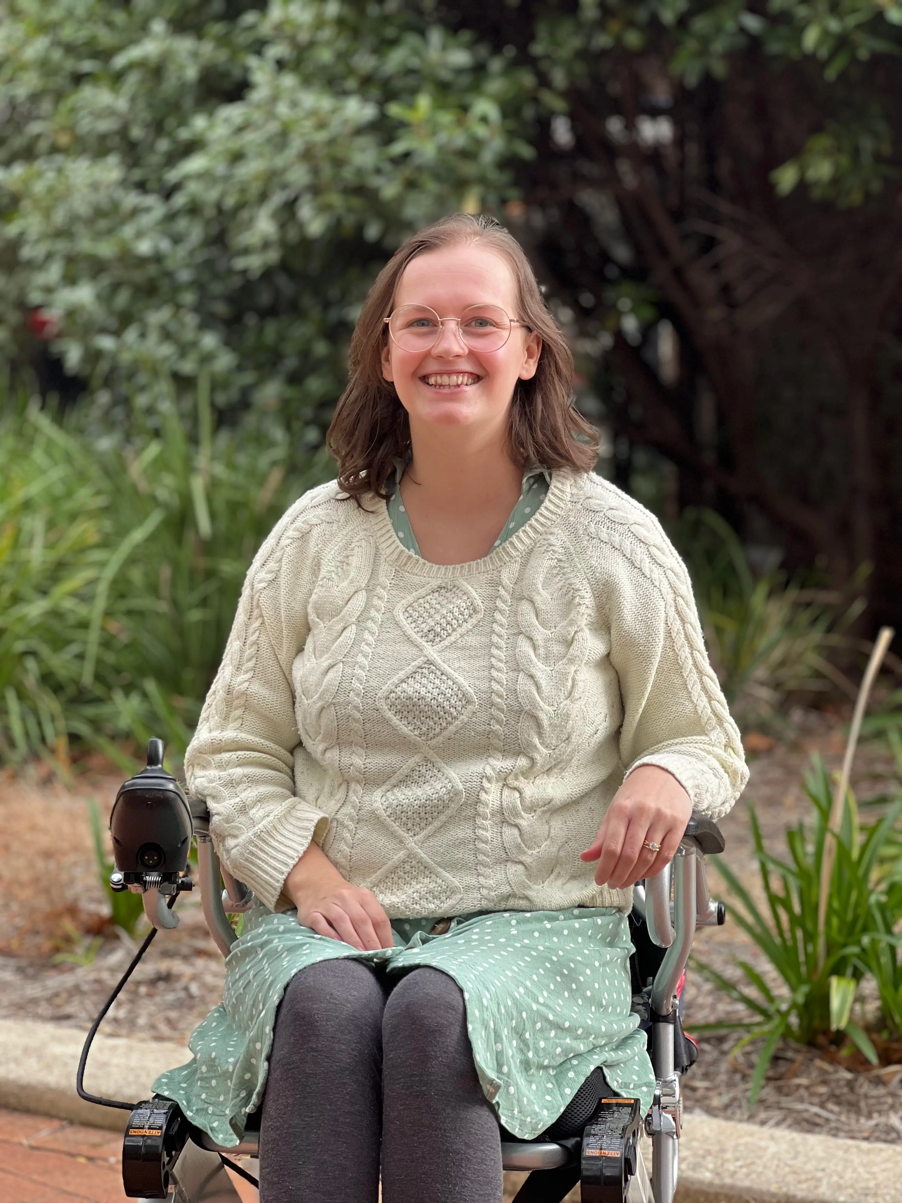 Cate sits smiling in a wheelchair in front of greenery. Her sage green dress has polka dots on it, and she wears an awesome knit sweater because the sun wasn't out for the photoshoot.