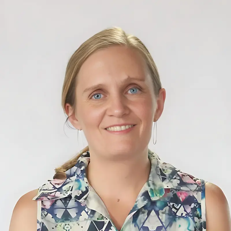 A woman with blonde hair pulled back smiles at the camera. She is wearing a sleeveless top with a geometric pattern and hoop earrings. The background is plain and light-colored.