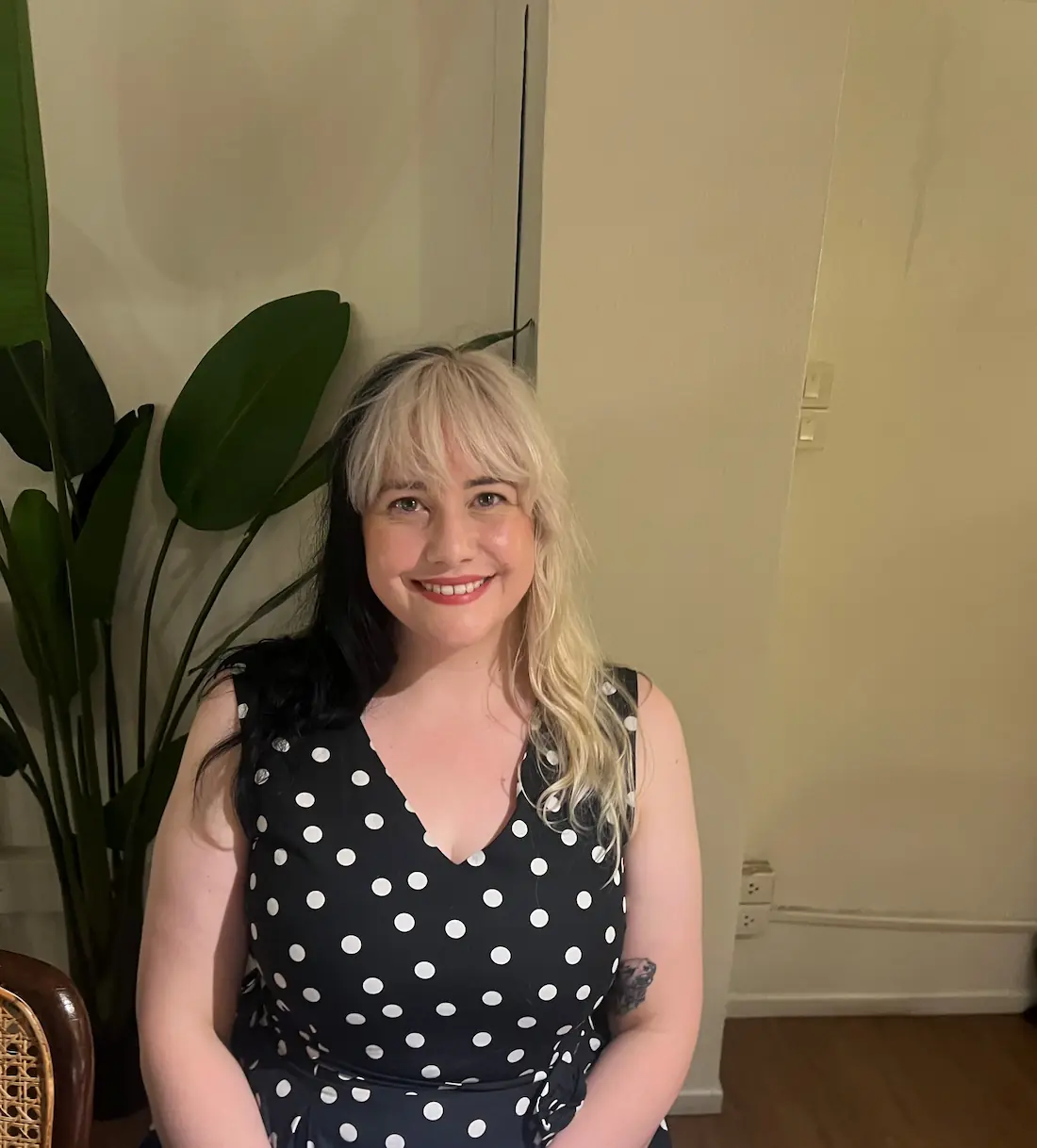 A smiling woman with split-dyed black and blonde hair, wearing a black sleeveless dress with white polka dots, seated indoors next to a large potted plant.