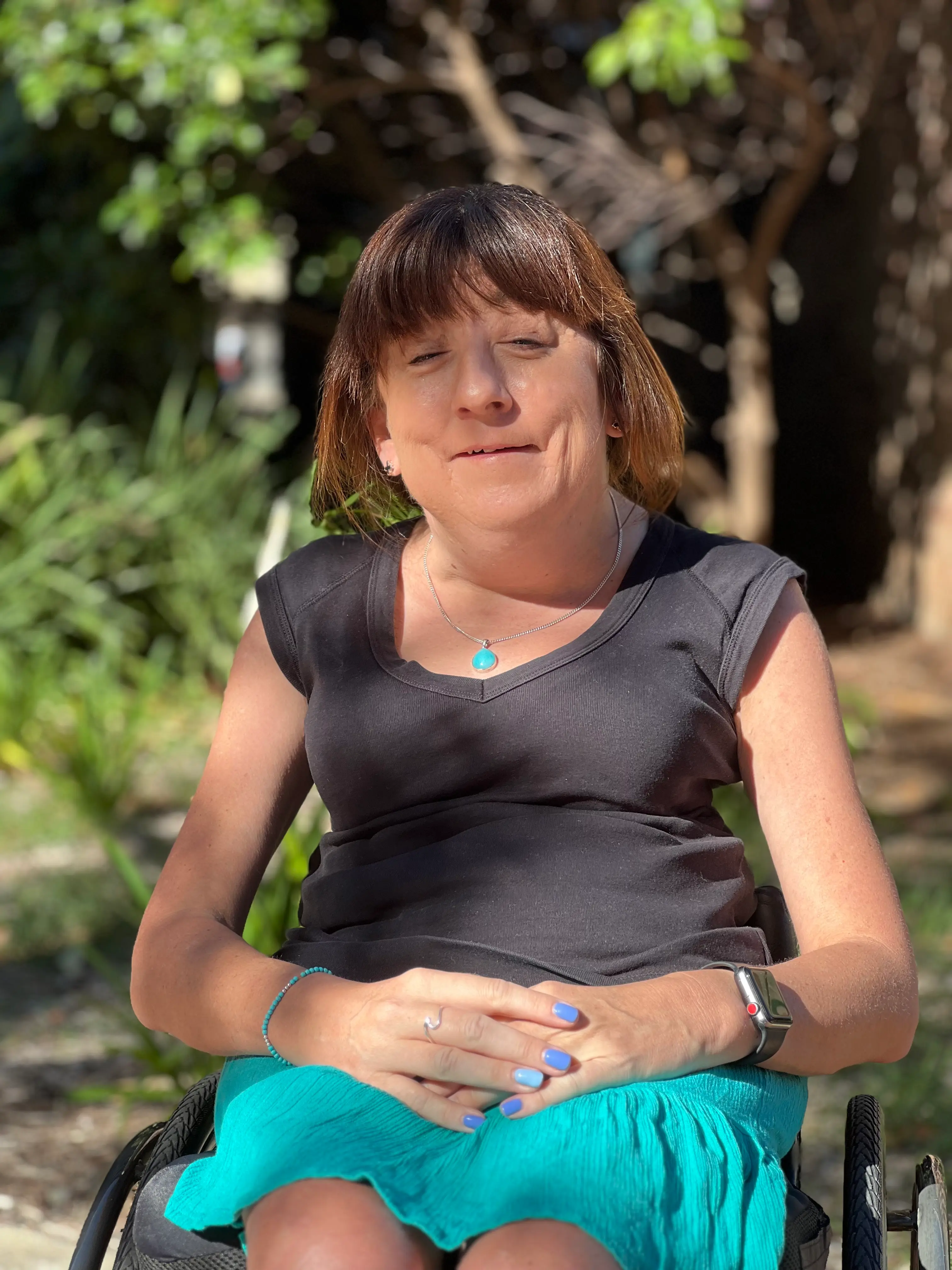 Mel sits smiling in her wheelchair in front of some nature in the background. She has paired her trademark blue nail polish with an aqua skirt (and necklace).