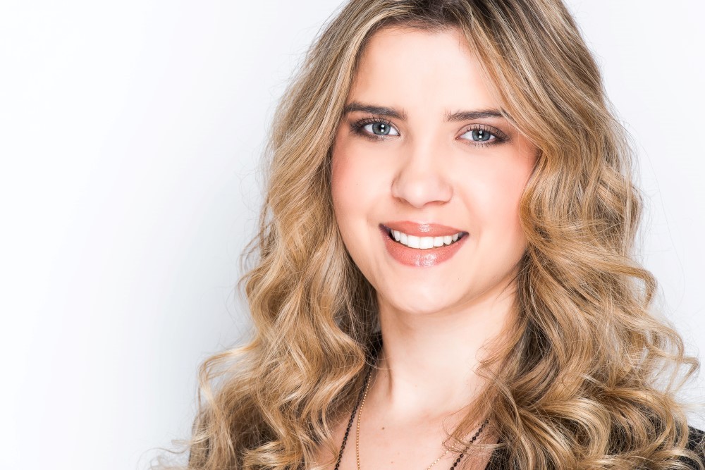 A woman with long, curly blonde hair smiles at the camera. She is wearing a black top and is posed against a plain white background.