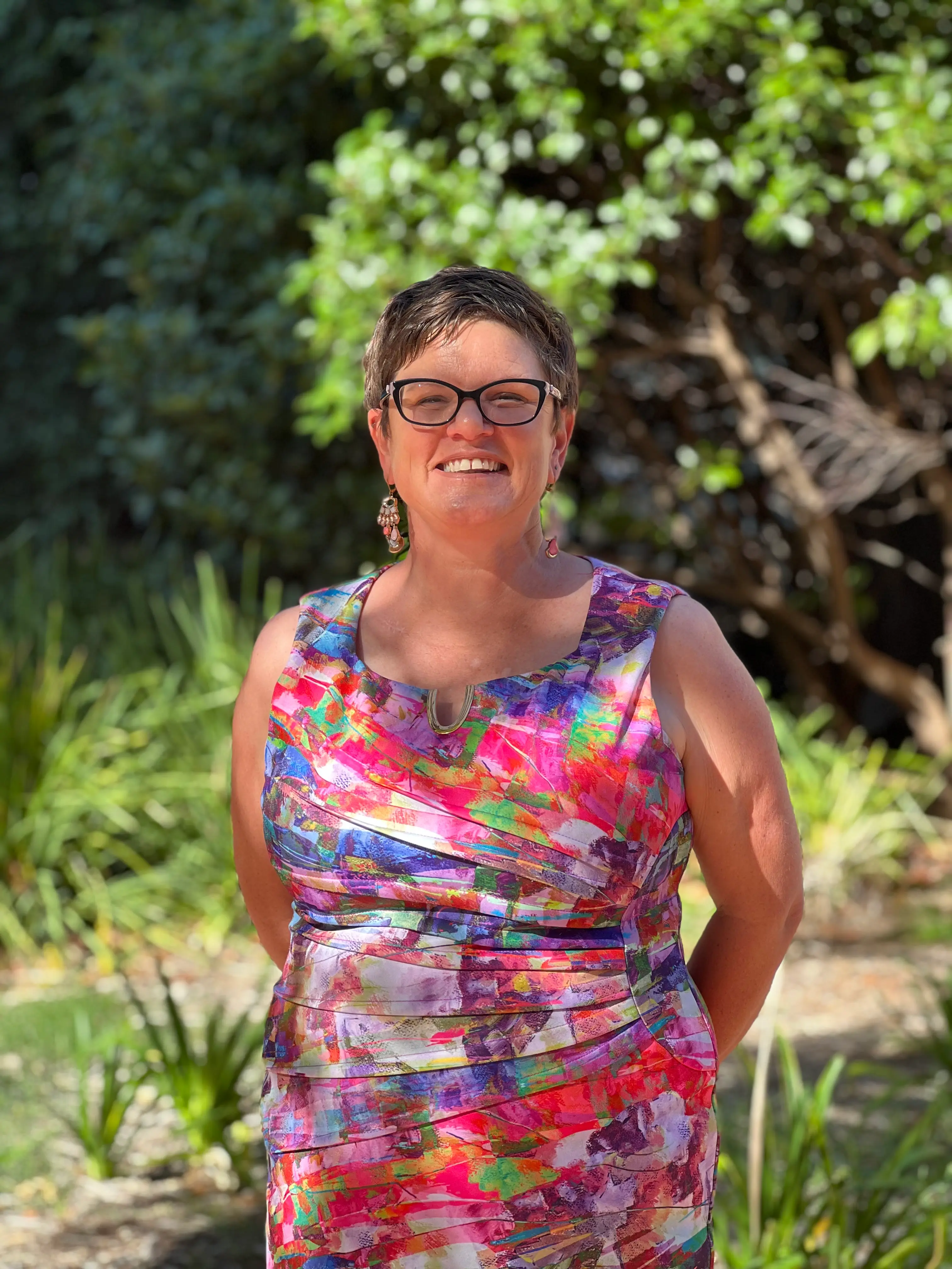 Sheridan stands smiling in front of greenery. Her multicolour dress pops against the background.