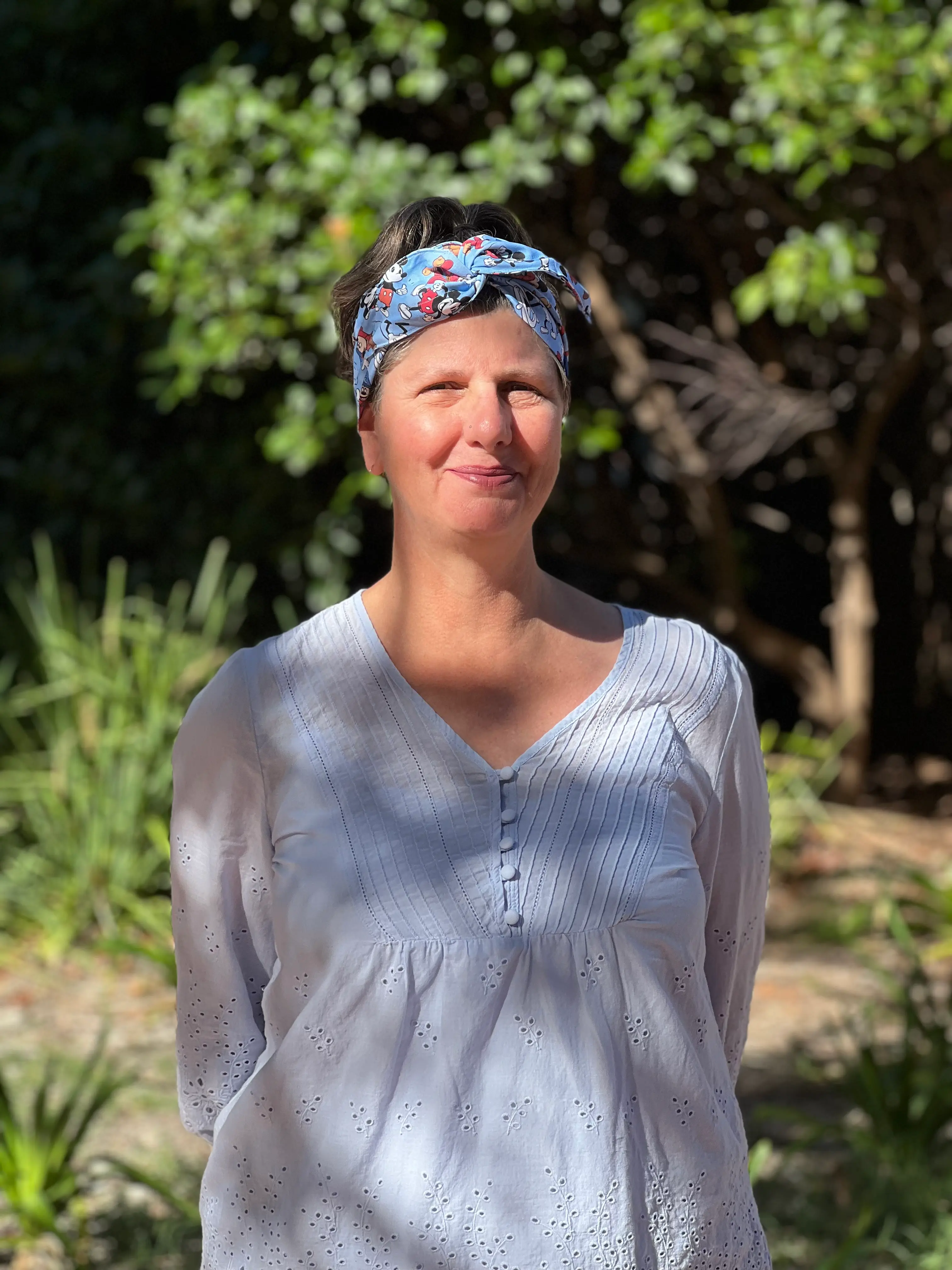 Sue stands smiling in front of greenery. Her pale blue shirt matches her Mickey Mouse hair wrap.