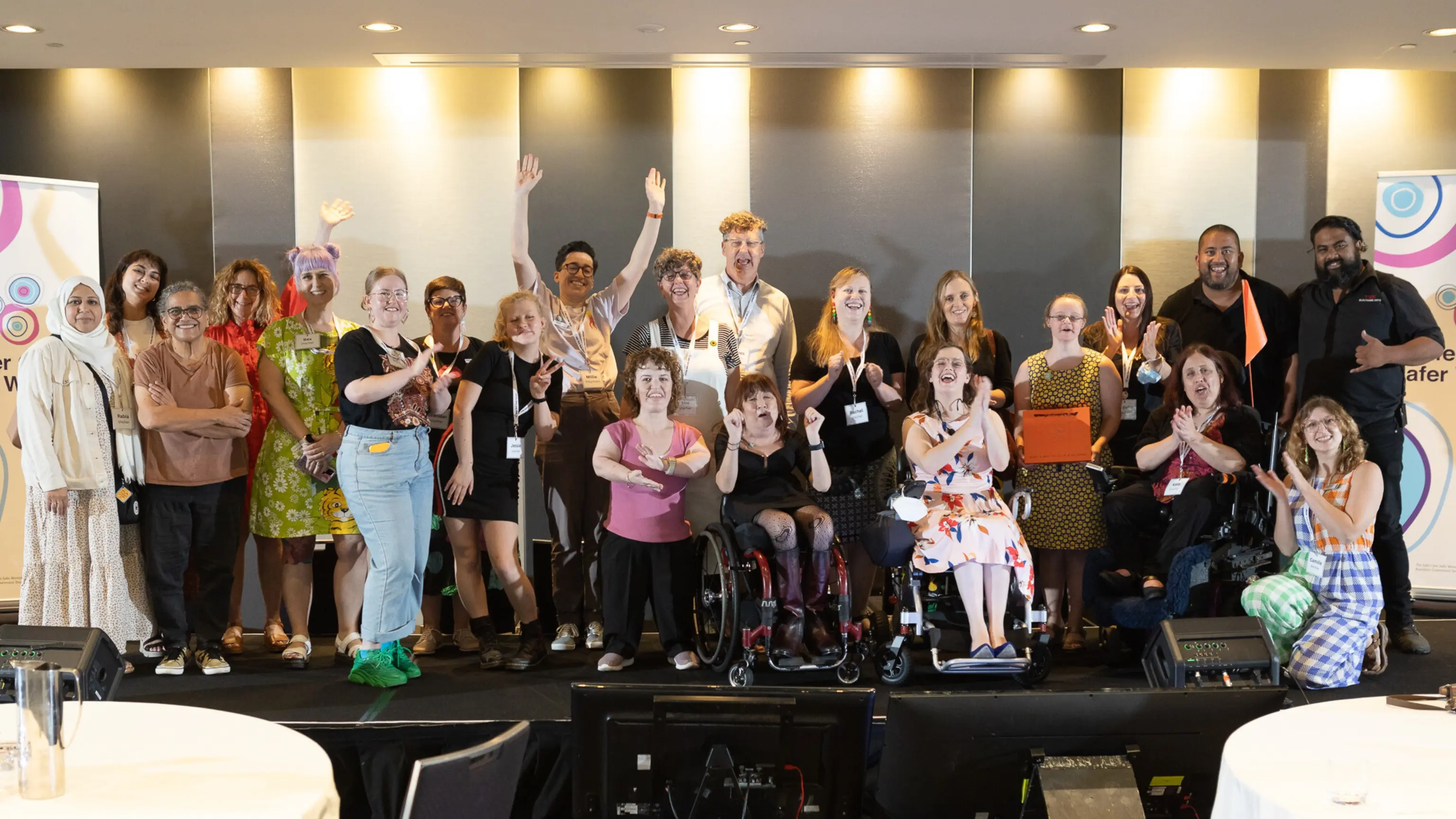 A diverse group of people poses together on a stage at a conference, smiling and making celebratory gestures. Some individuals are standing, while others are seated in wheelchairs. The background features two banners with colorful designs, one on each side of the group.