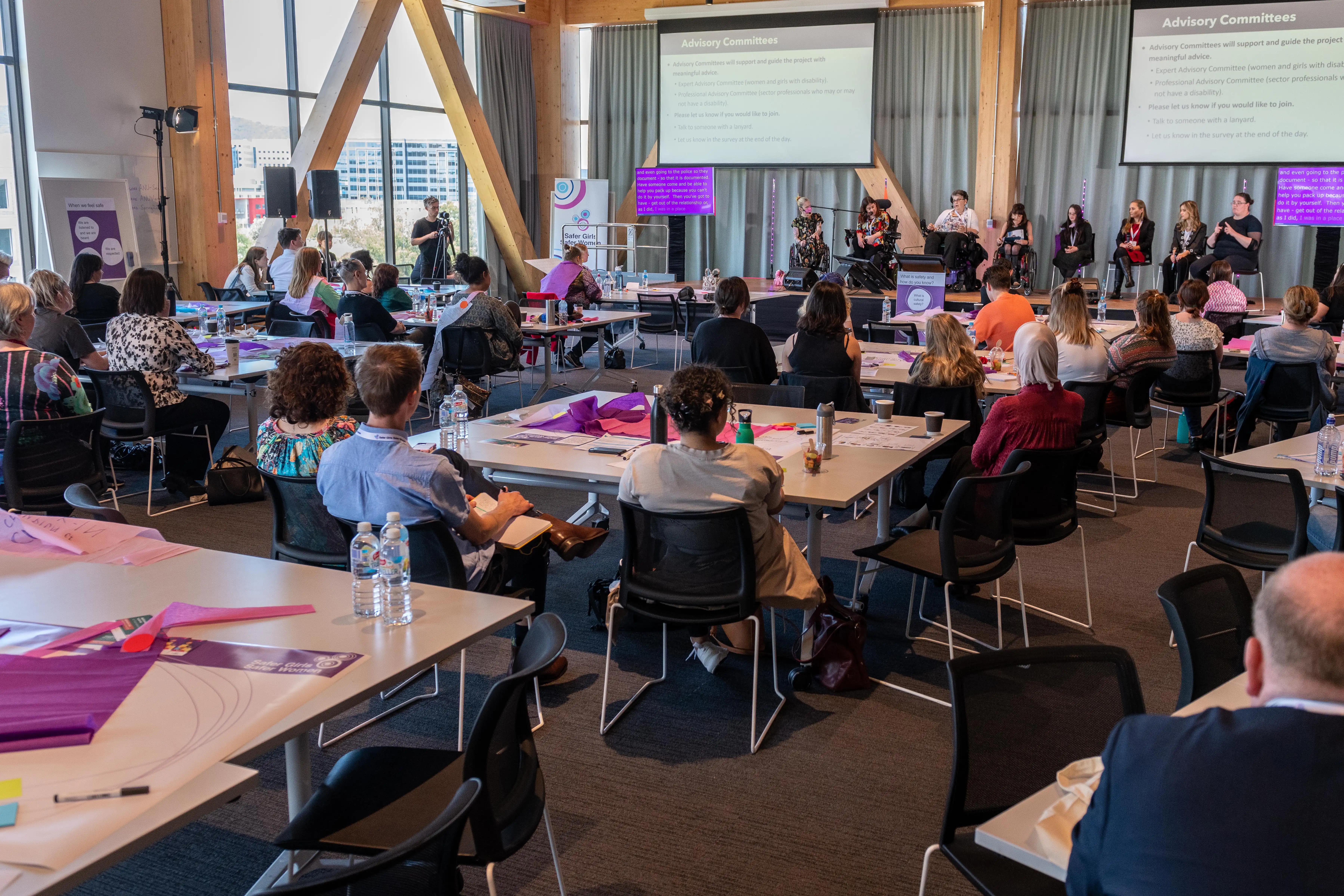 A conference setting with attendees seated at tables, facing a stage with speakers and presenters. Two large screens display presentation slides titled Advisory Committees. Attendees are engaged, with some taking notes and others listening attentively.