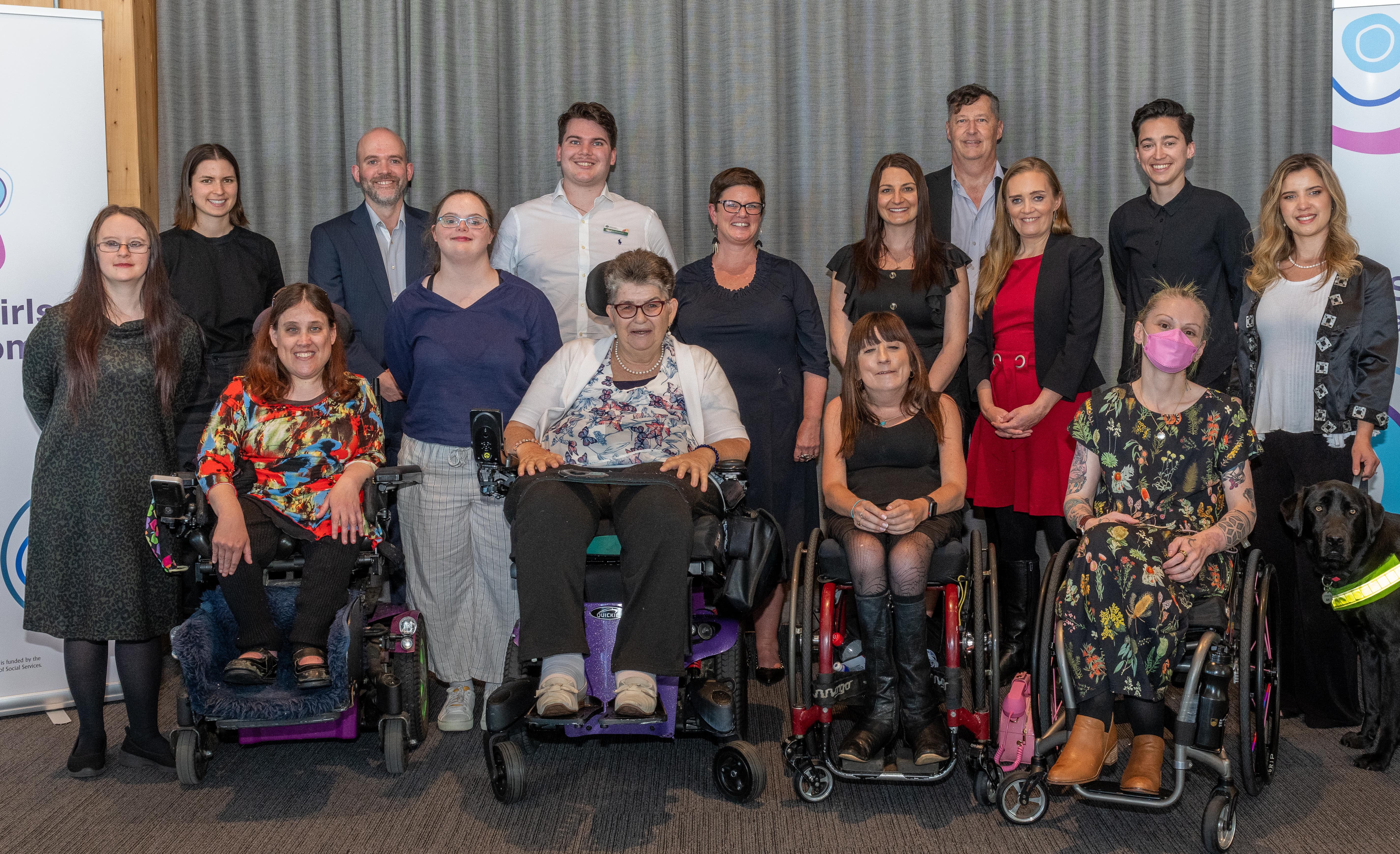 A group of people poses together, some standing and some seated in wheelchairs, in front of a gray curtain.