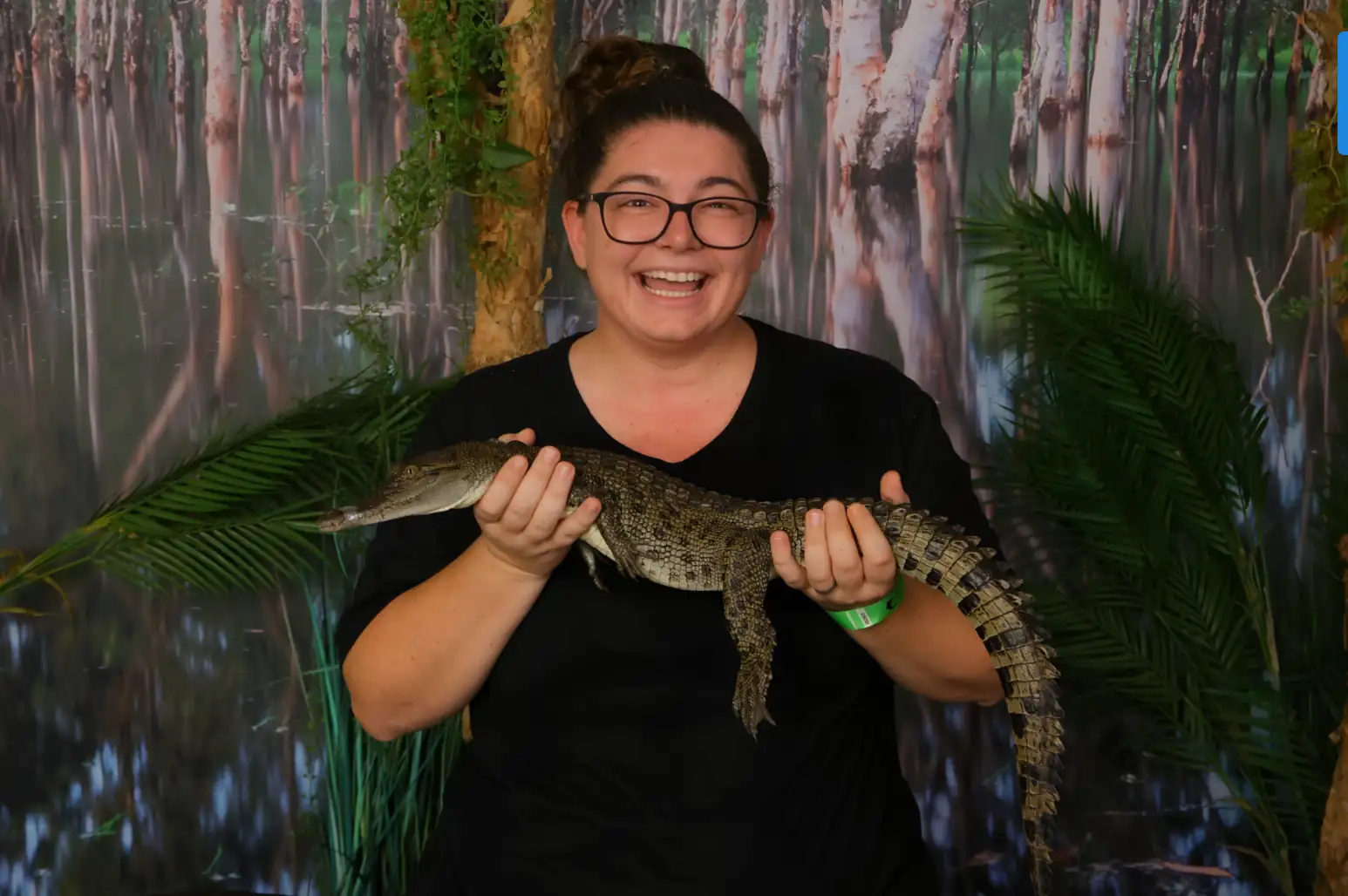A smiling woman wearing glasses and a black shirt holds a small juvenile crocodile with both hands against a backdrop of a swampy forest.