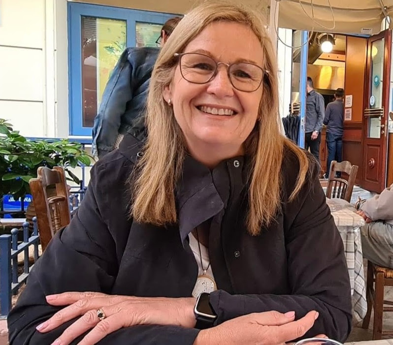 A woman with blonde hair and glasses smiles while sitting at a table in an outdoor café. She is wearing a dark jacket, and there are people and a colorful storefront in the background.