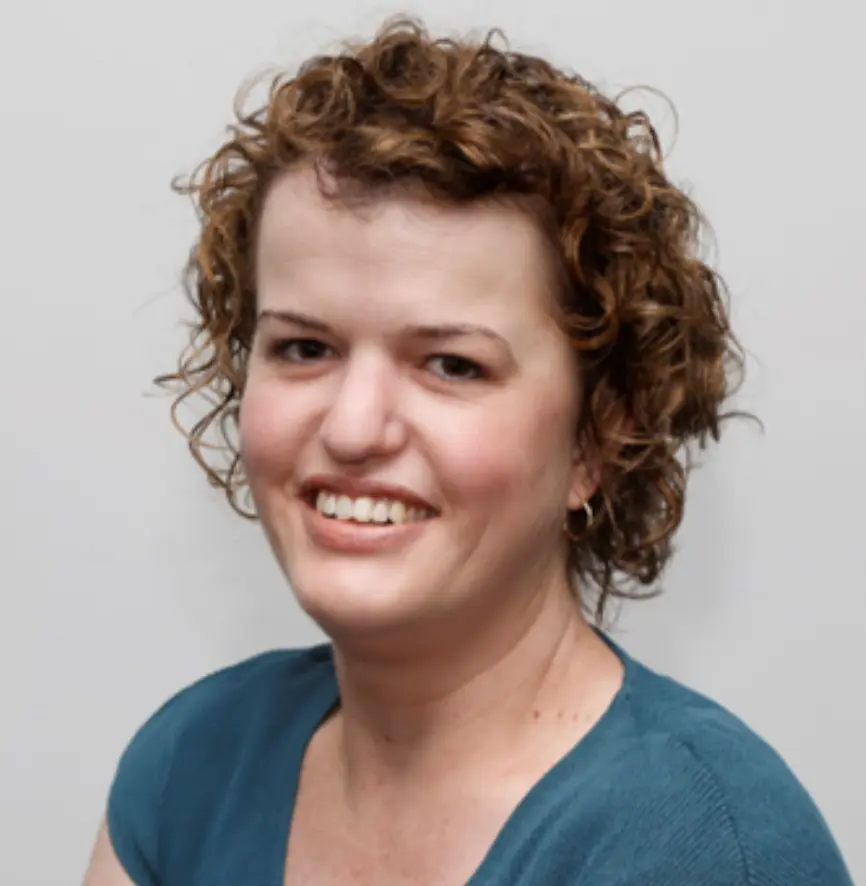 A woman with short curly brown hair, wearing a teal top, smiles at the camera. She wears small hoop earrings.