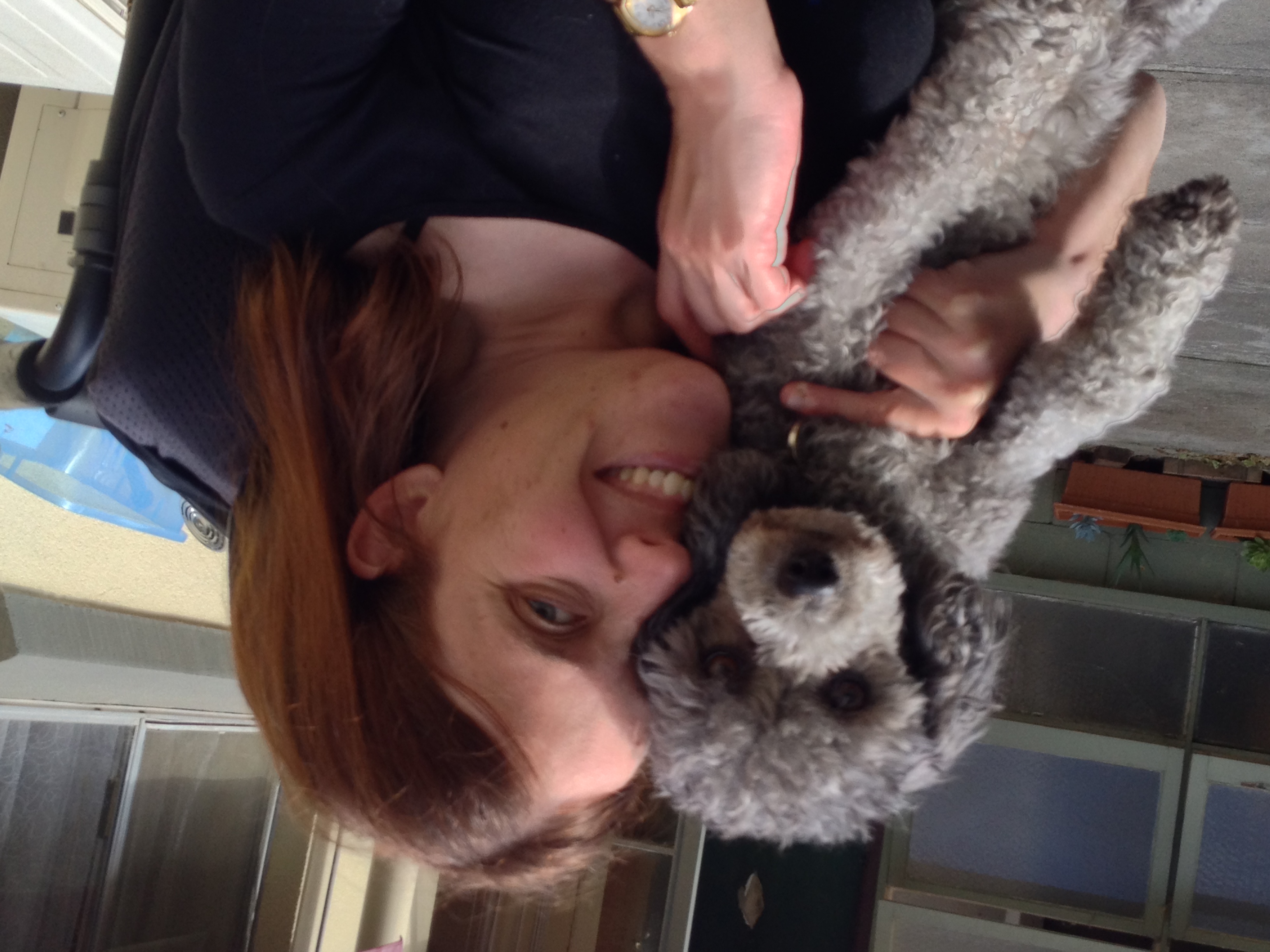 A woman with brown hair smiles while holding a gray poodle. They are outside in a patio area with potted plants and windows in the background.