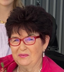 A woman with short dark hair and red glasses smiles while wearing a red top and pearl earrings. She is in an indoor setting with part of another person visible behind her.