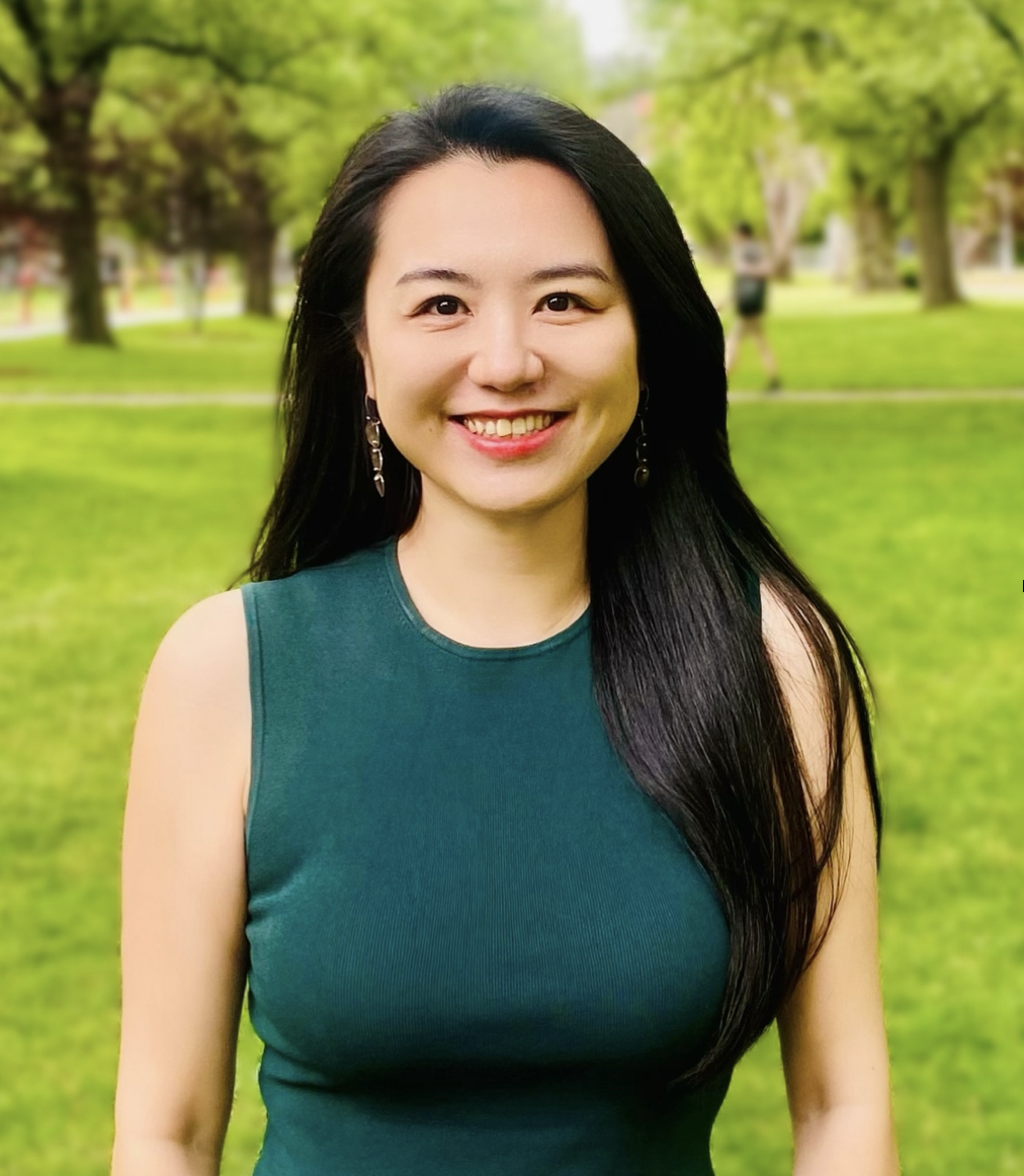 A woman with long black hair and wearing a sleeveless green top smiles at the camera while standing in a park with green grass and trees in the background.