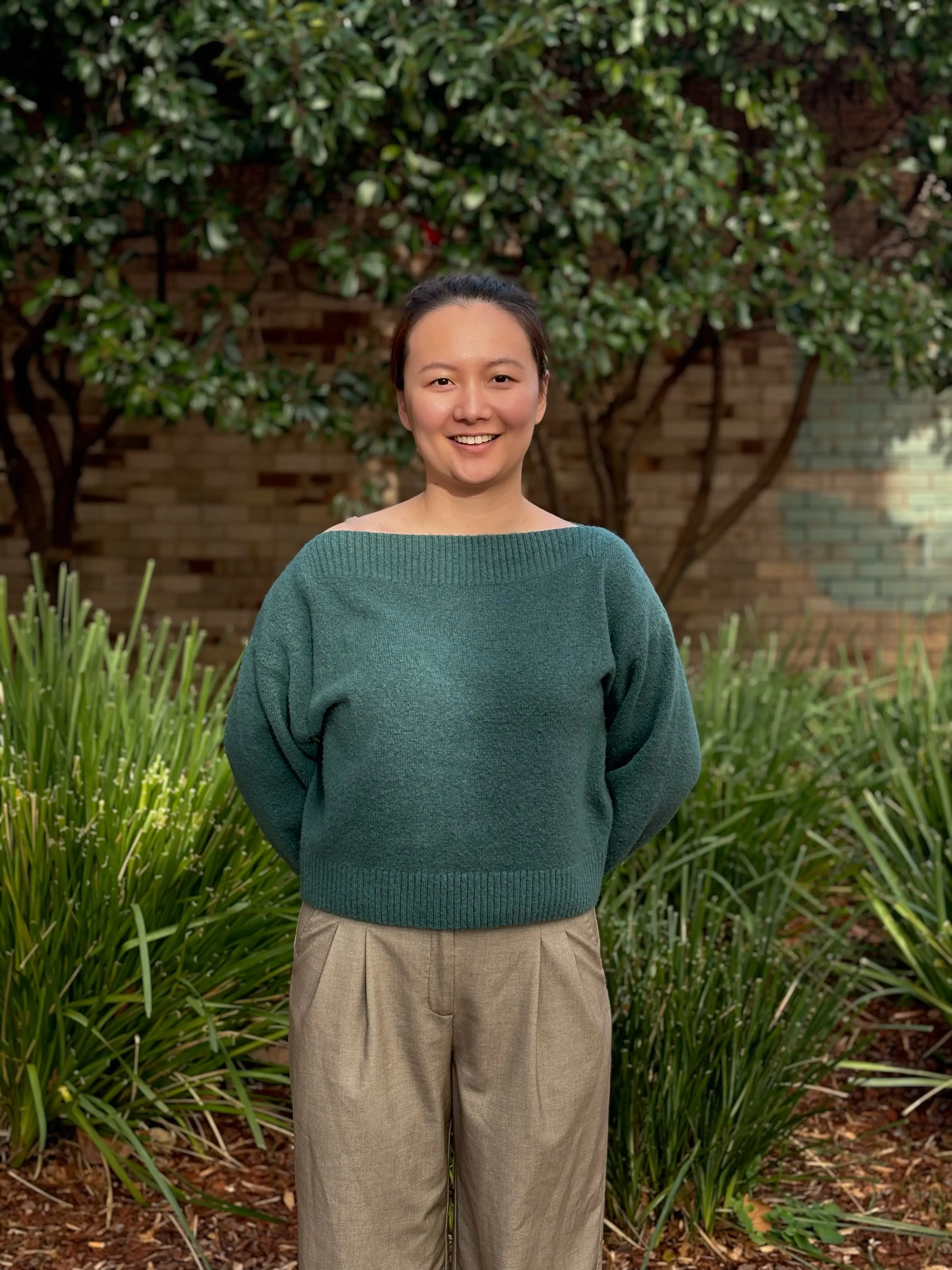 Maggie stands smiling in front of greenery in a forest green sweater and light brown pants.