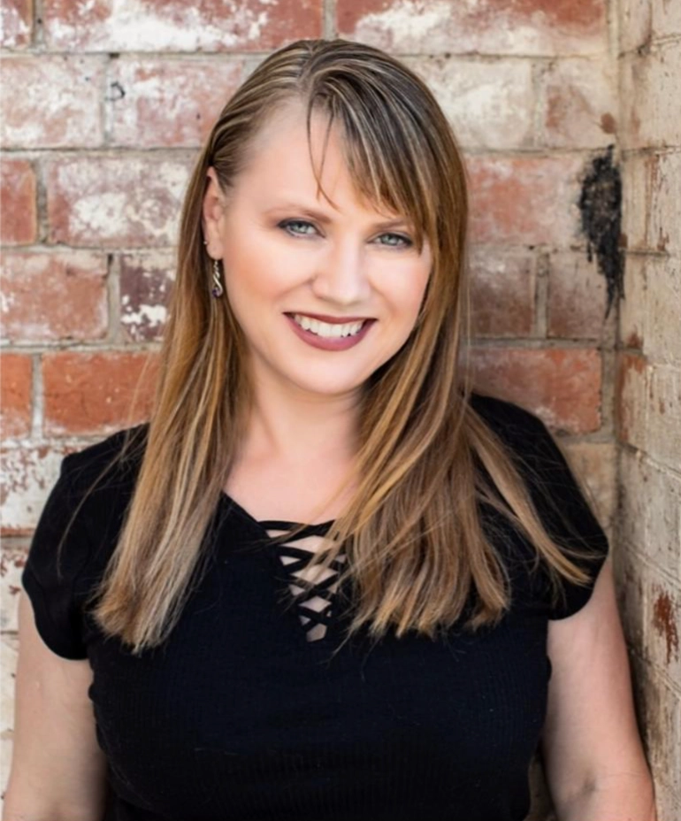 A woman with straight, light brown hair and side-swept bangs smiles while standing in front of a brick wall. She is wearing a black top with a crisscross design at the neckline.