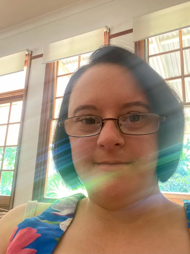 A woman with short dark hair and glasses takes a selfie indoors. She is wearing a colorful top, and sunlight is streaming through the windows behind her, creating a lens flare effect.