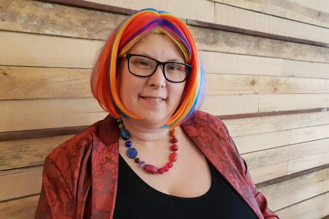 Portrait of Yenn with colorful rainbow hair, glasses, and a red patterned jacket, against a wooden background.